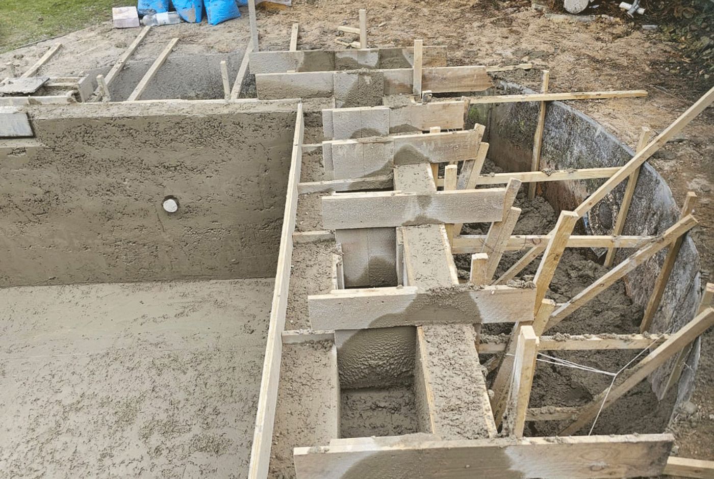 One corner of a concrete swimming pool shell with wooden supports and exposed soil around.