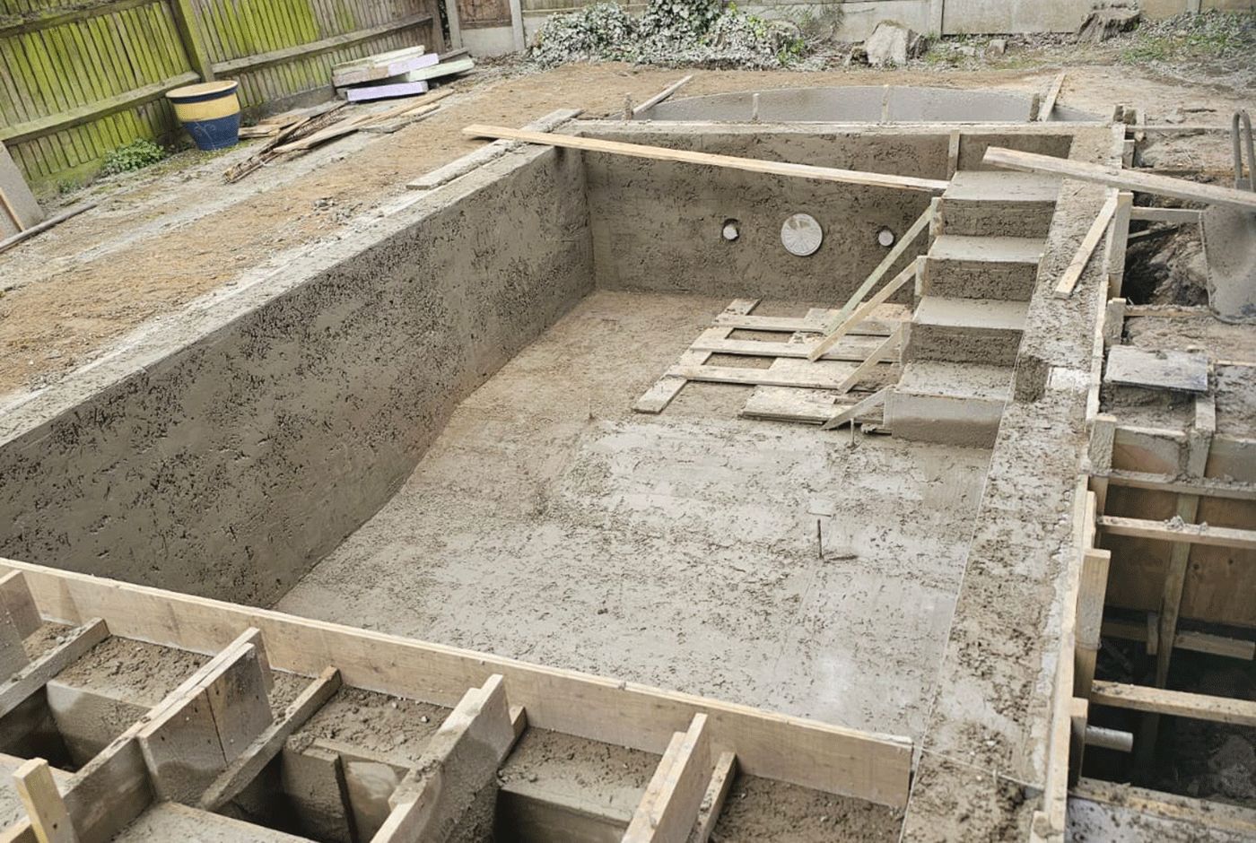 Concrete shell of a swimming pool under construction with steps down and wooden formwork visible.