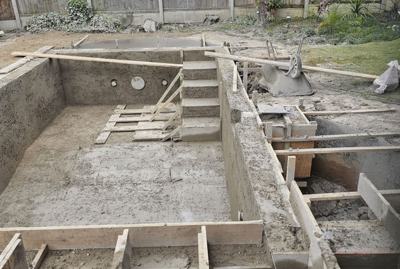 Concrete swimming pool shell with steps under construction in a residential garden.