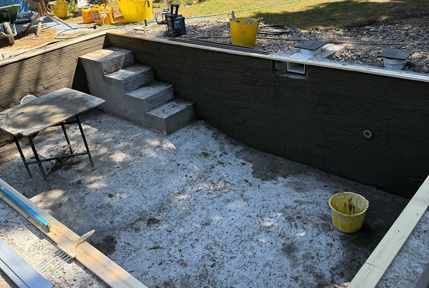 Concrete shell of a small pool in a garden, workers' tools and equipment in and around the shell.