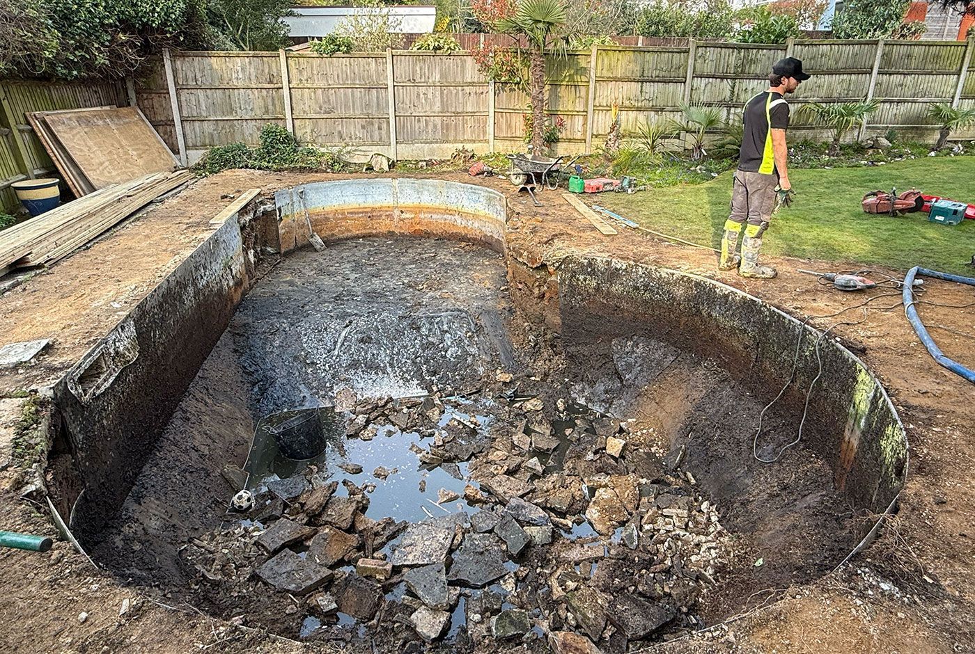 A small swimming pool in a garden being removed, mud and rubble in its base, fencing and materials in the background.