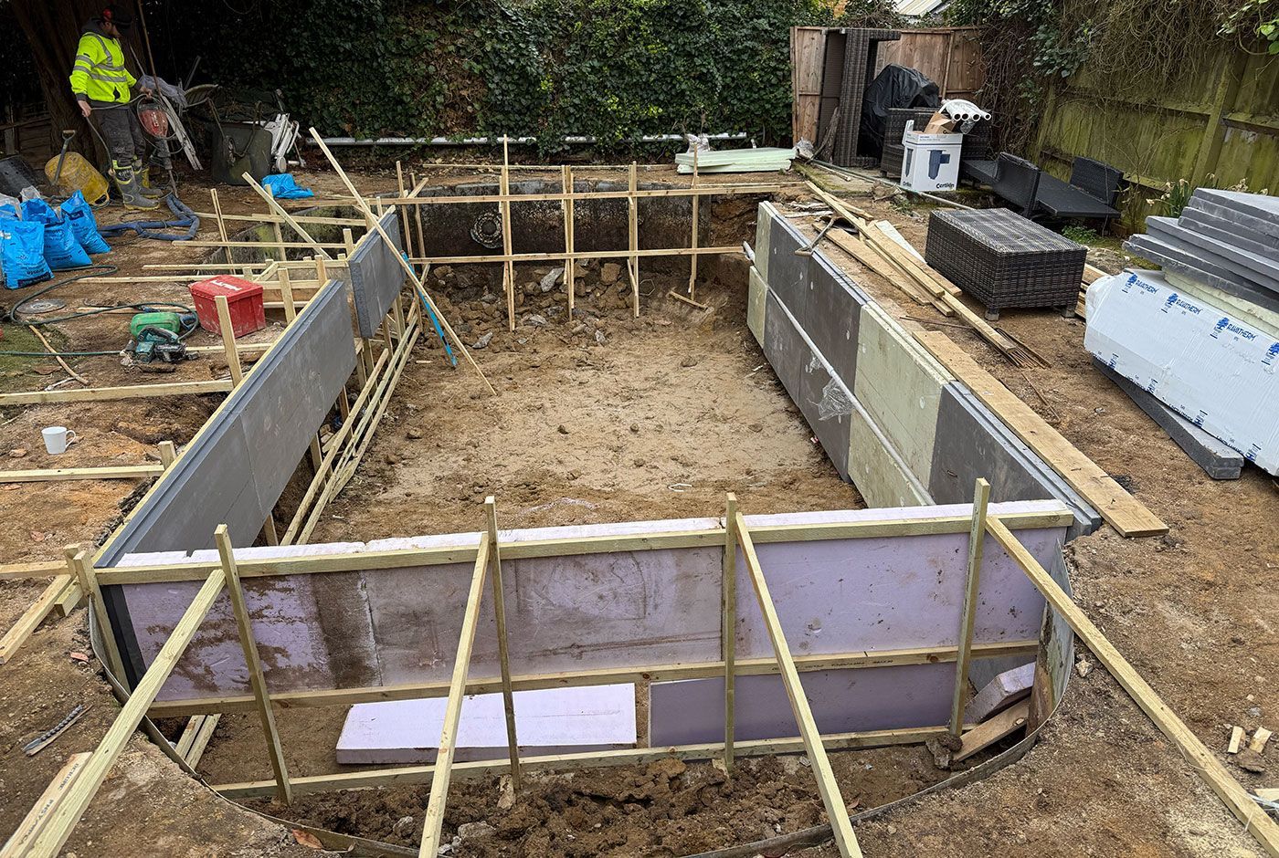 Construction site of a garden swimming pool with a large rectangular wooden foundation form and workers in the background.