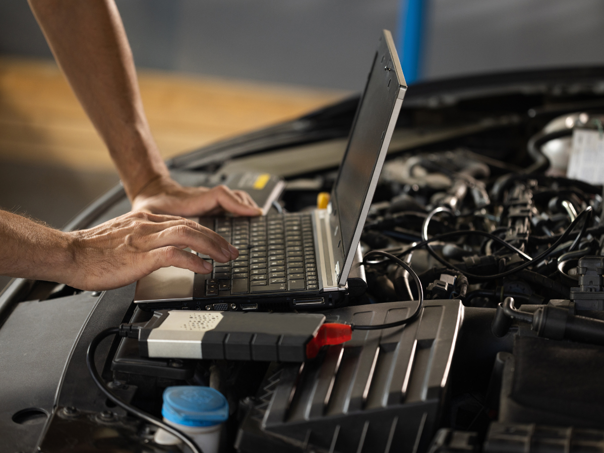 A mechanic using a laptop and diagnostic device to inspect a car engine in a garage.