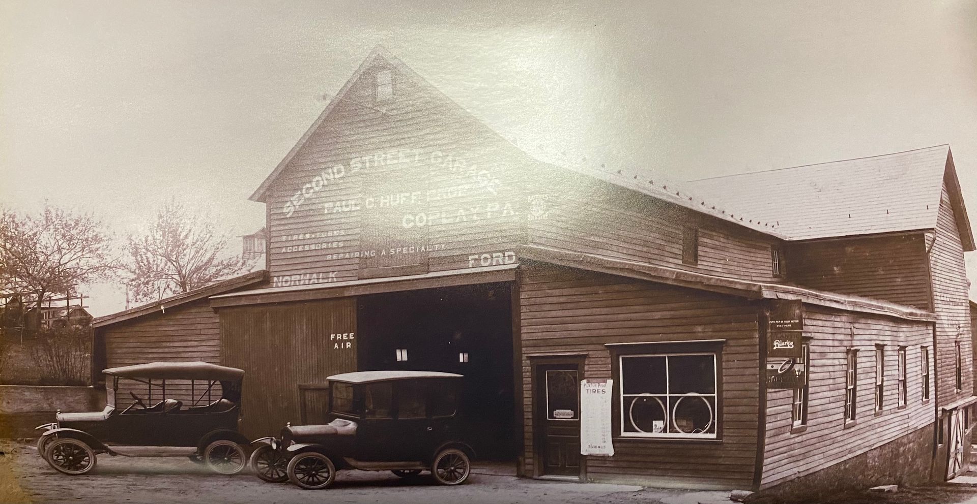 Sepia-toned old wooden garage with vintage cars parked outside on a dirt lot | Herstine Automotive LLC