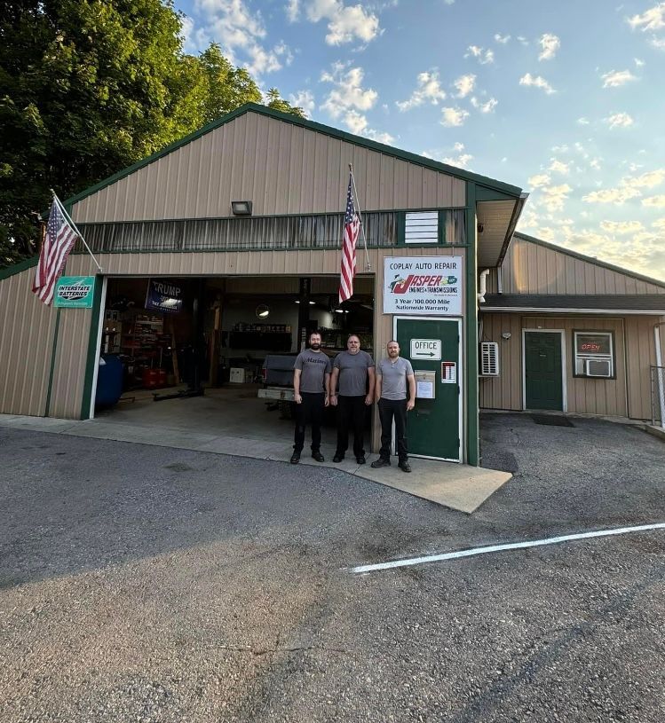 Three people standing outside a tan garage-style building with American flags at sunset | Herstine Automotive LLC