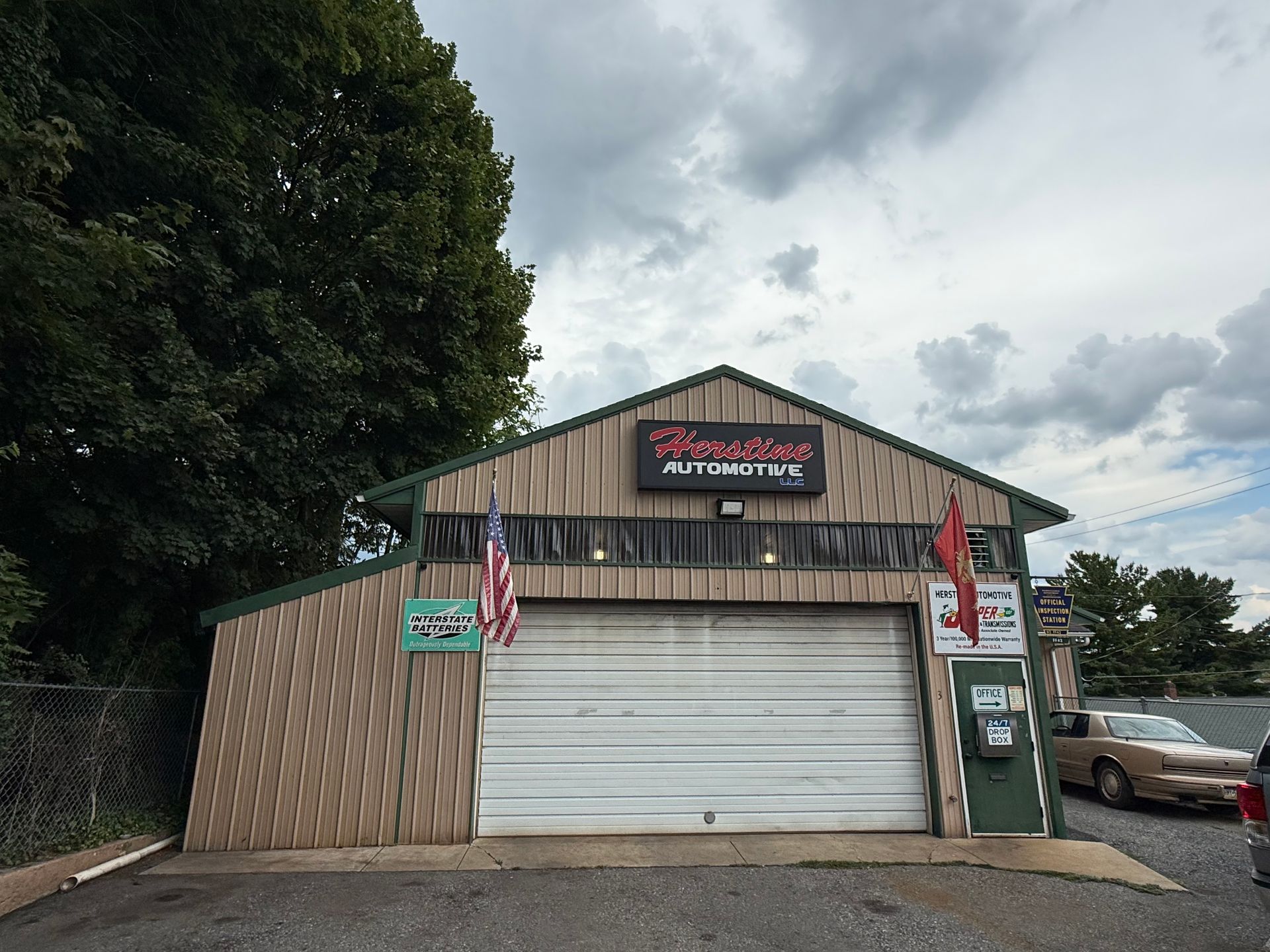 Small rustic roadside shop with a white garage door, American flags, and parked cars under cloudy skies. | Herstine Automotive LLC
