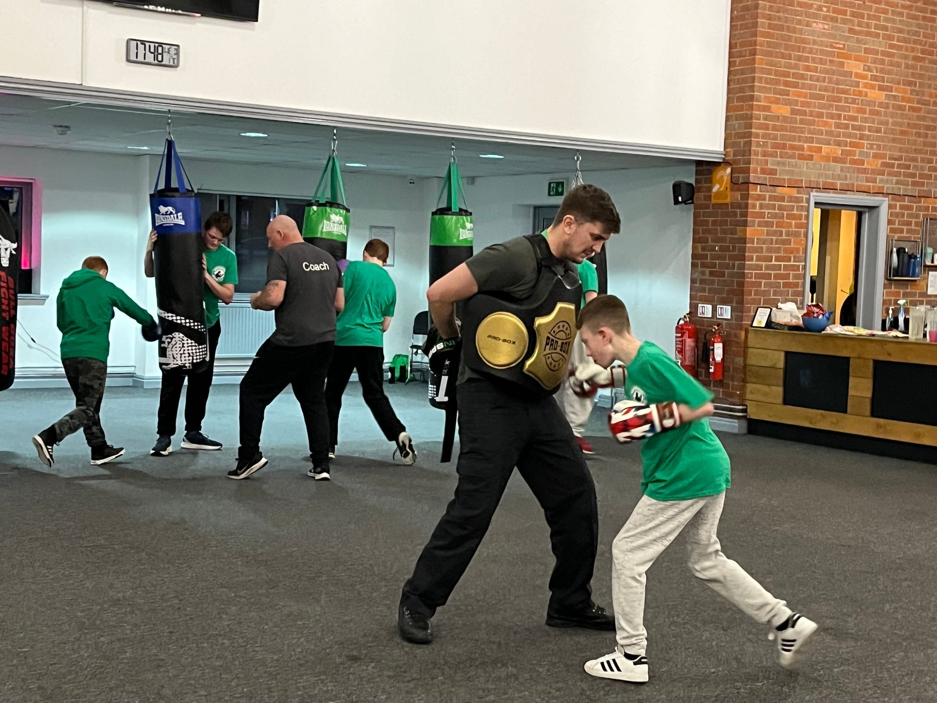 Boxing training: Young boy wearing gloves punches as man holds focus mitt. Gym setting, others boxing.
