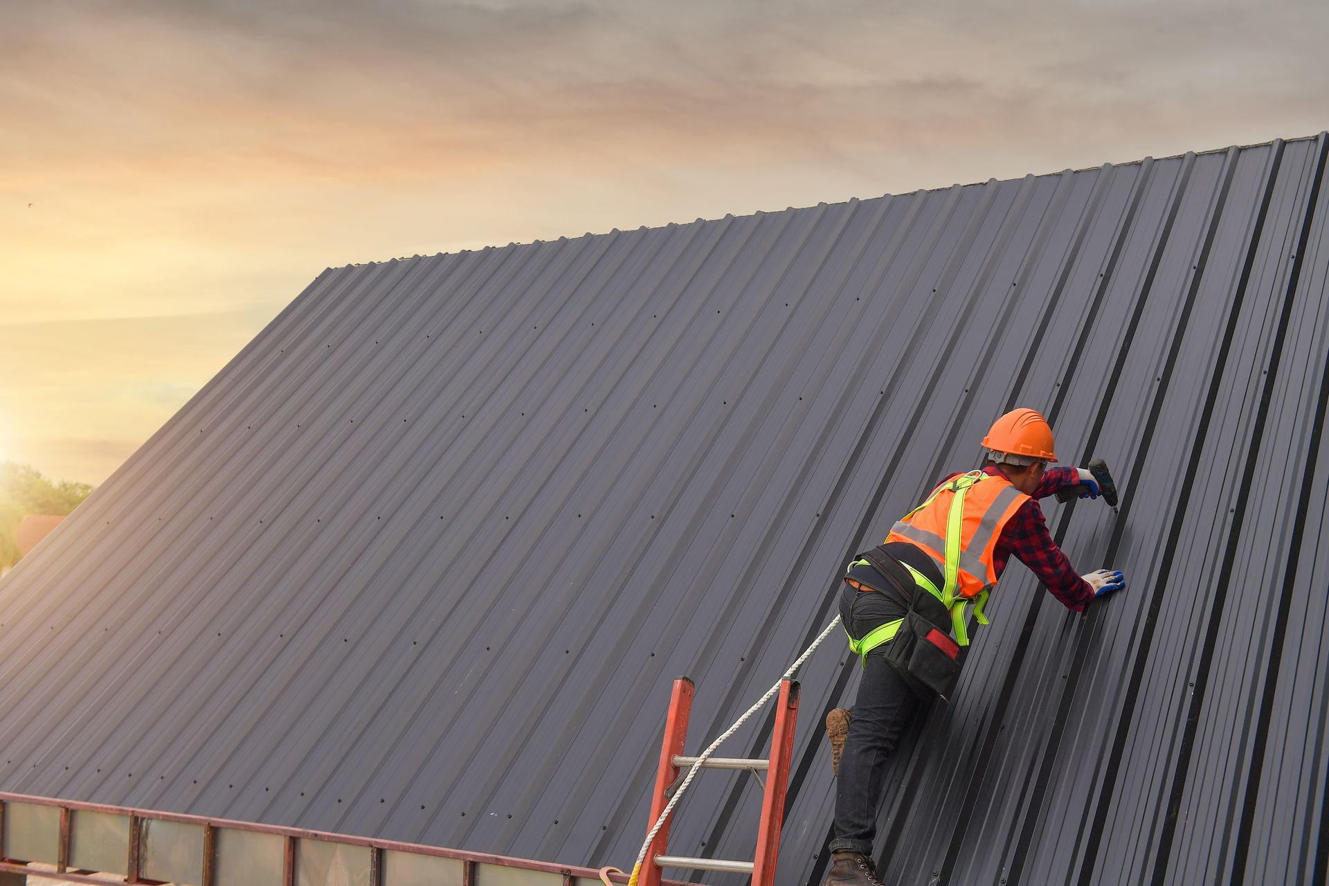 Construction worker in safety gear installing dark metal panels on a sloped roof at sunset.