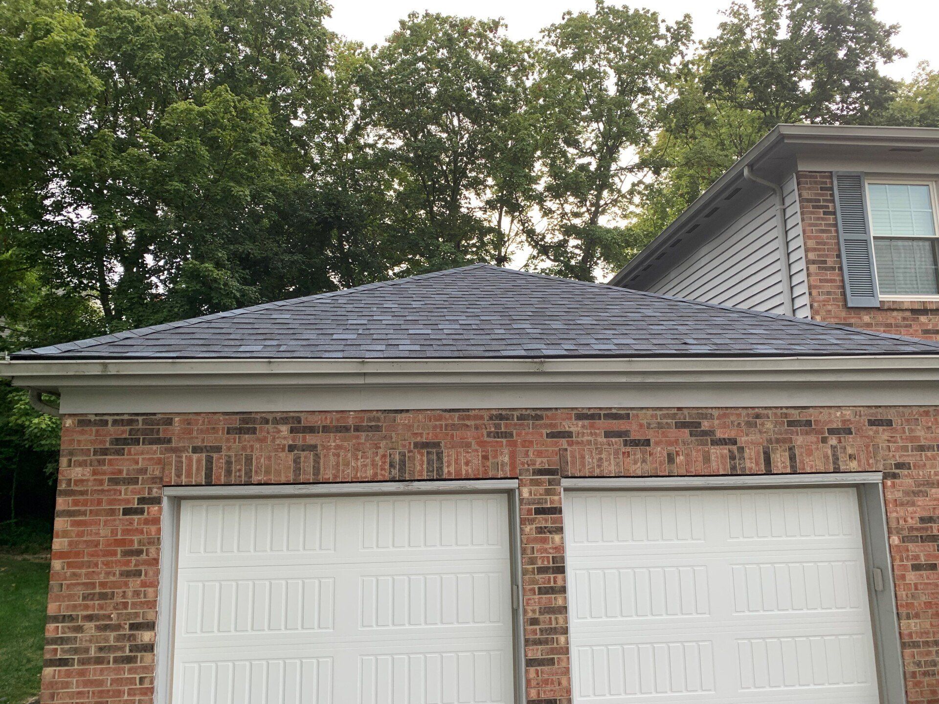 Brick garage with white doors, dark roof, and trees in the background.