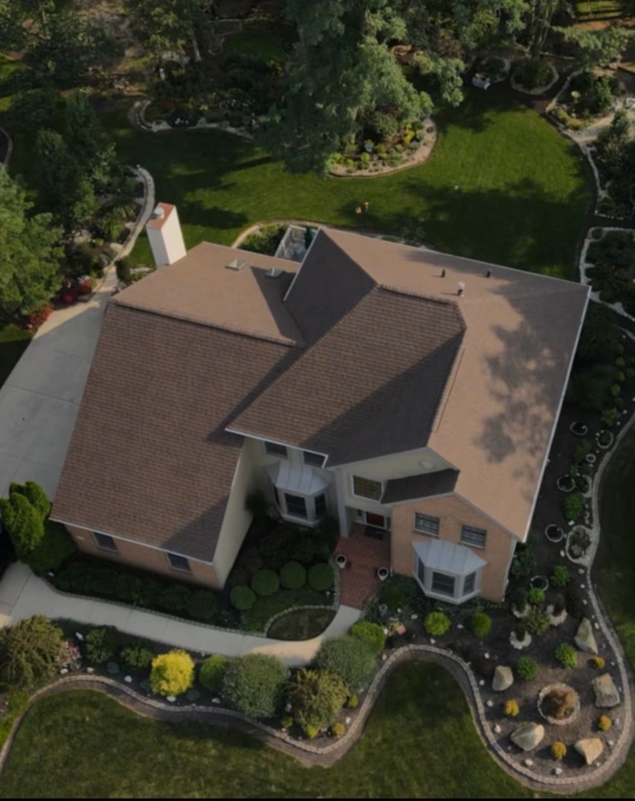 Aerial view of a large house with brown roof, surrounded by green lawns, trees, and landscaping.