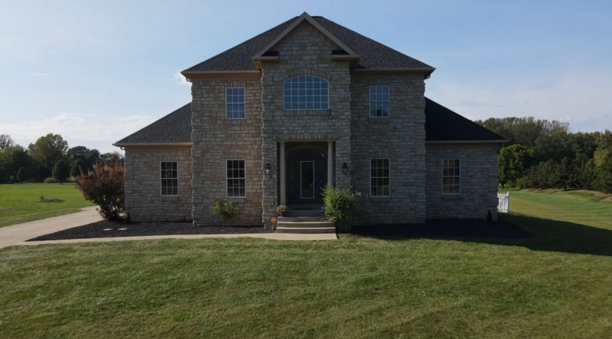 Two-story brick house with dark roof and large front windows, set on a green lawn on a sunny day.