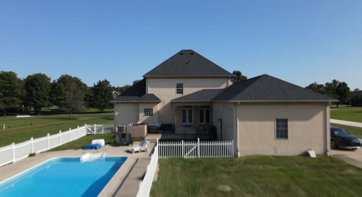 Back of a two-story beige house with a pool, white fence, and black roof, set against a sunny sky and green lawn.