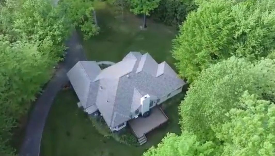 Overhead view of a house with a gray roof, surrounded by green trees and a paved driveway.