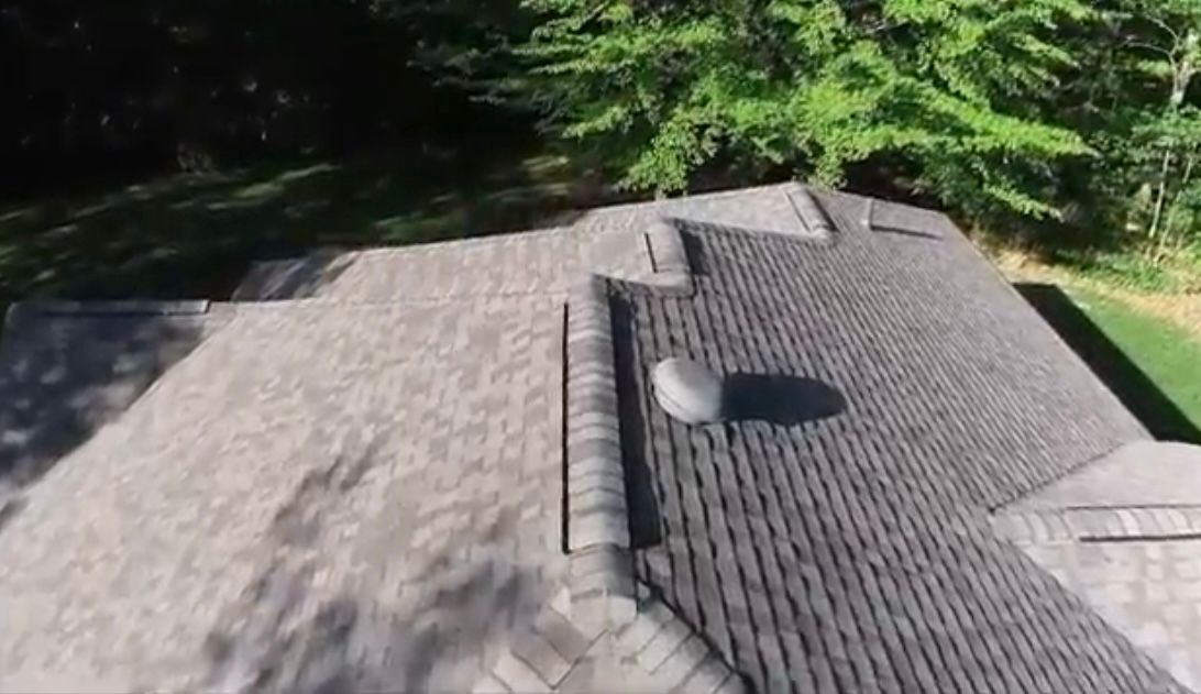 Overhead view of a house roof with shingles. The roof is surrounded by trees and grass.