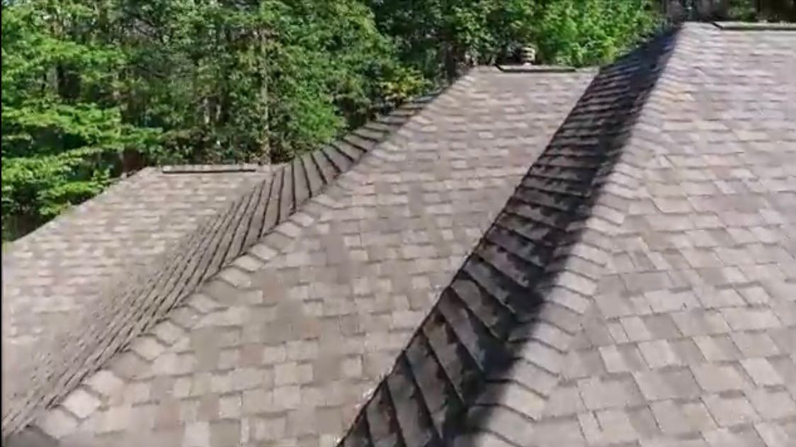Overhead view of a house roof with brown asphalt shingles and a background of green trees.