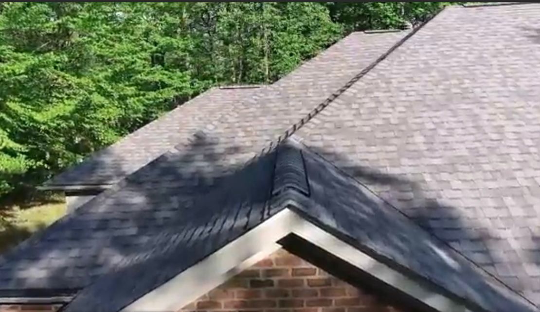 Overhead view of a roof with brown asphalt shingles and a brick facade, trees in the background.