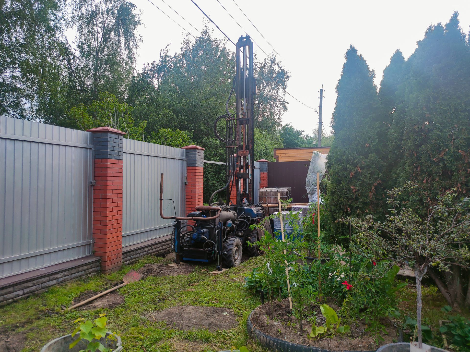 A residential well drilling rig set up in a backyard garden beside a brick and metal fence