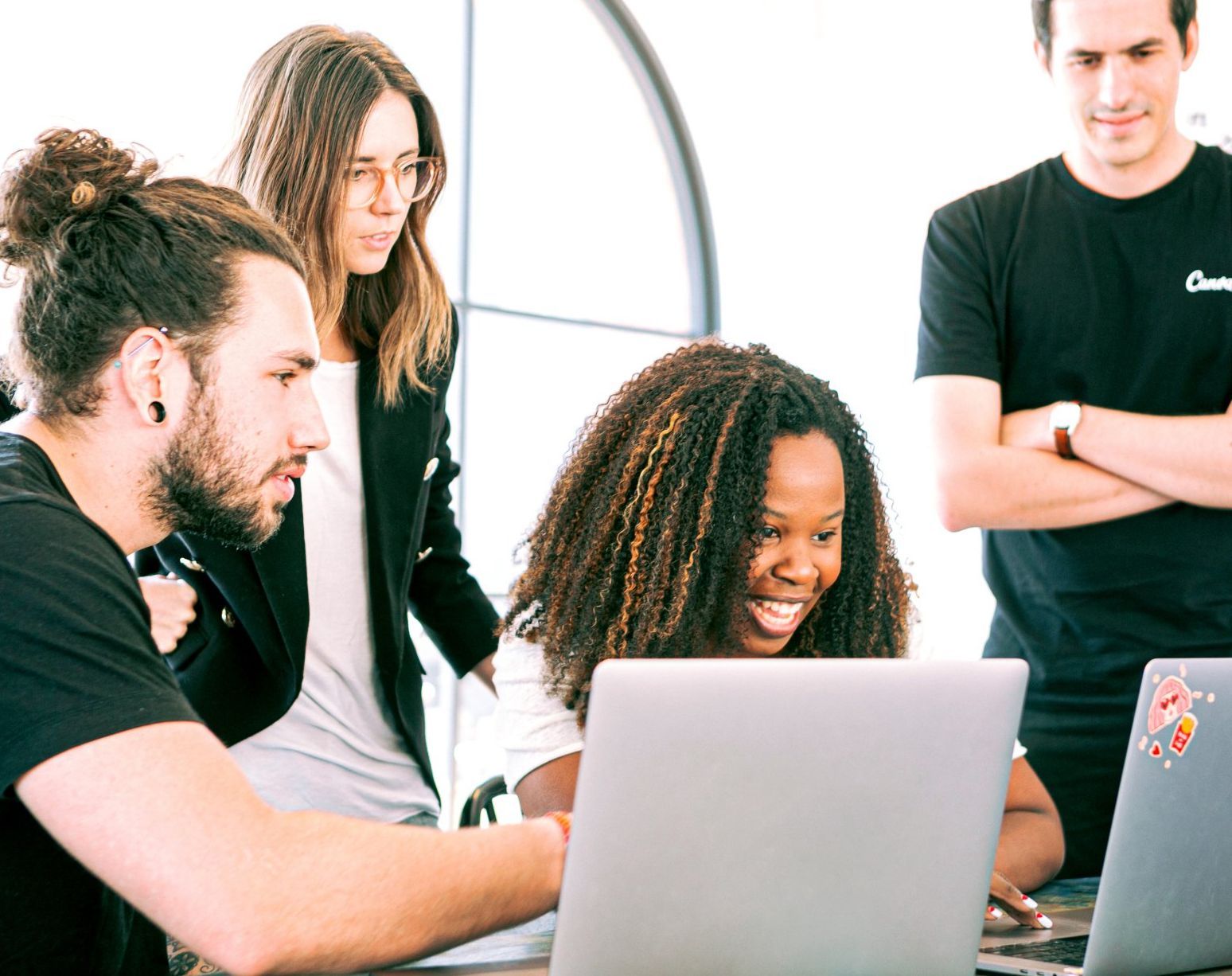Four people looking at a laptop, one smiling. Modern office setting with natural light.