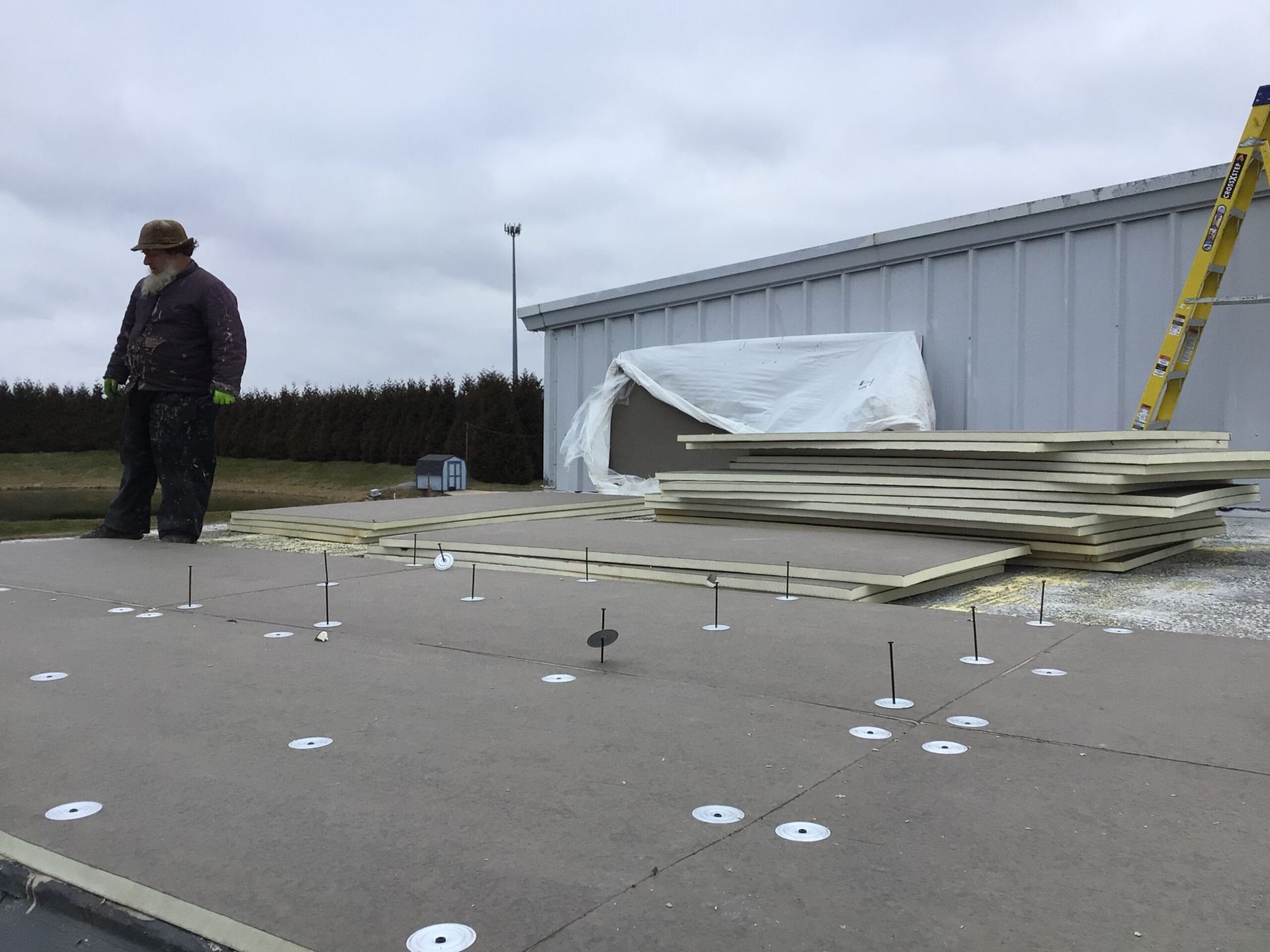 Man standing on flat roof, installing insulation panels. Building in background, cloudy sky.
