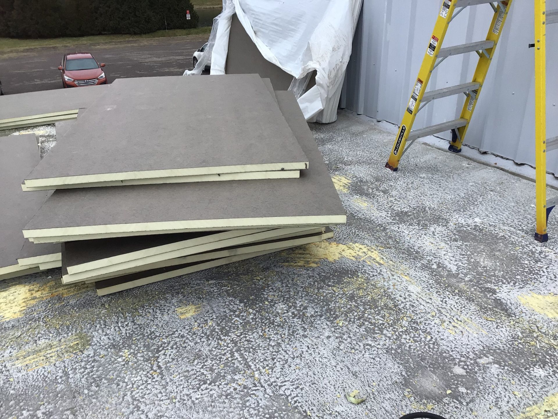 Pile of gray insulation panels on a concrete surface, a yellow ladder leans against a building.