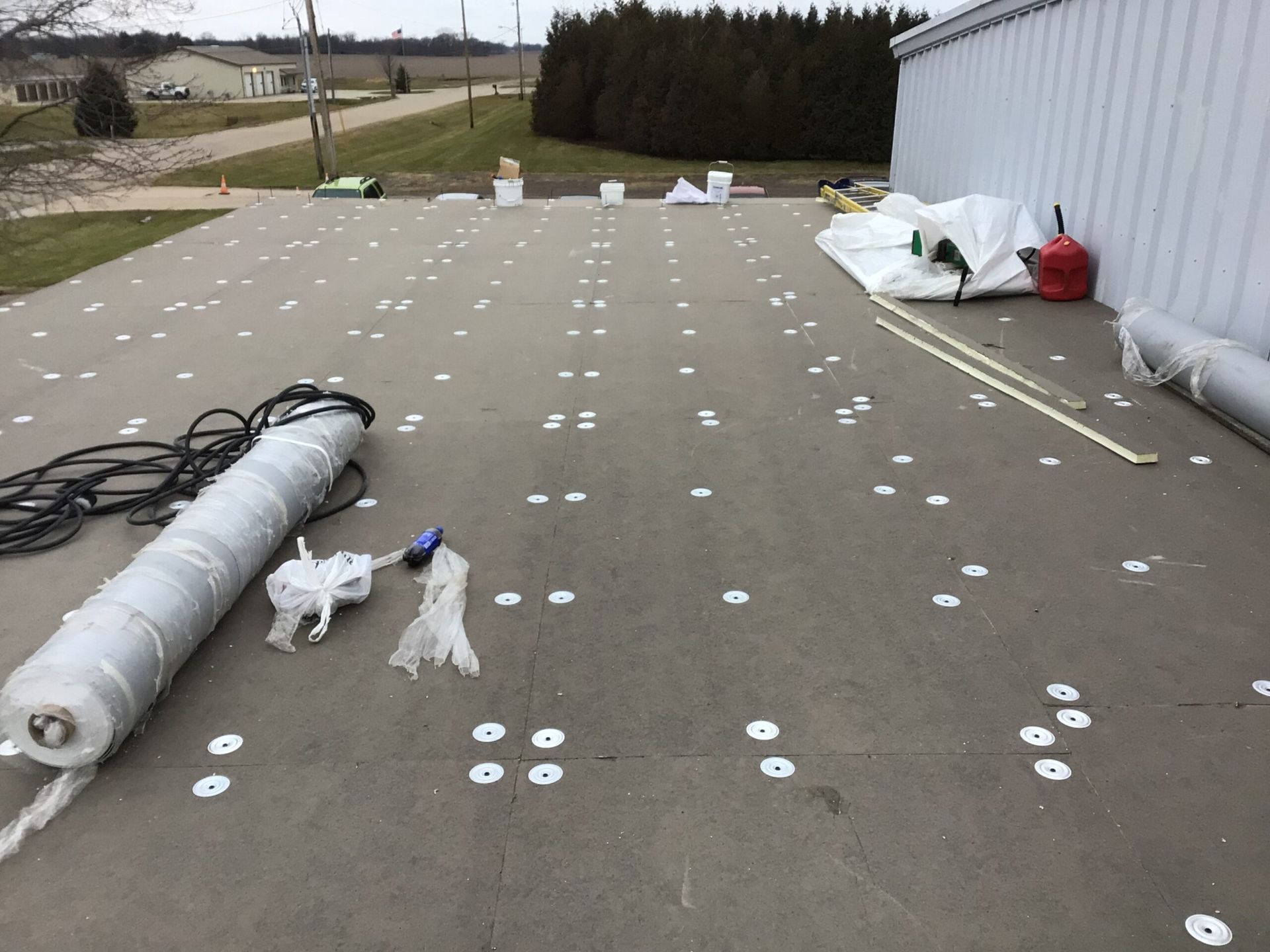Flat roof with numerous white fasteners; rolls of material, tools, and a propane tank sit nearby.