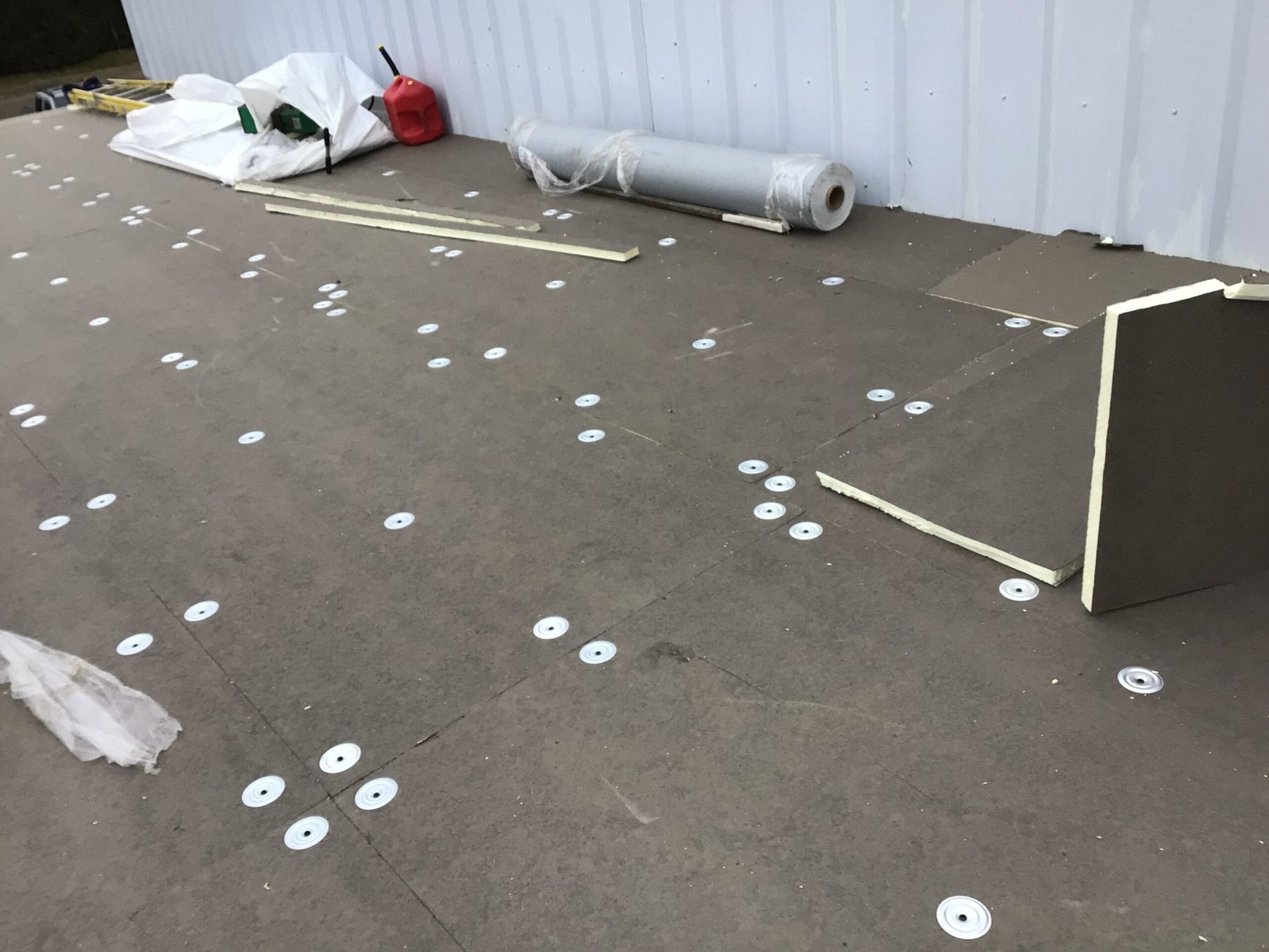 Flat roof with dark panels, white fasteners, and construction materials like rolls and bags against a white building.