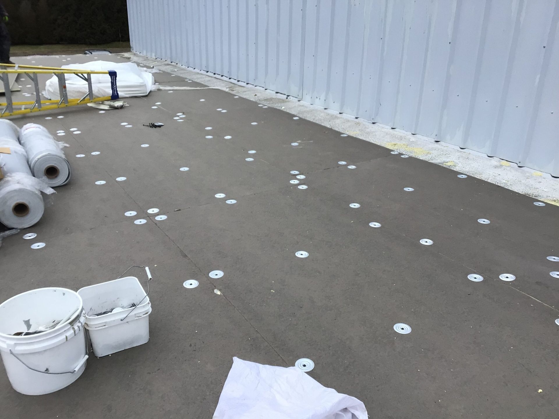 A roofing project in progress. Gray surface with numerous white washers scattered around buckets and rolls of material.