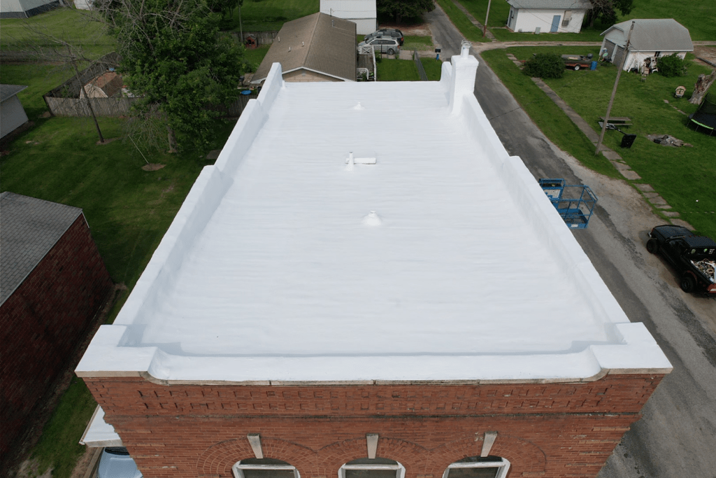Overhead view of a brick building with a white flat roof, surrounded by green grass and streets.
