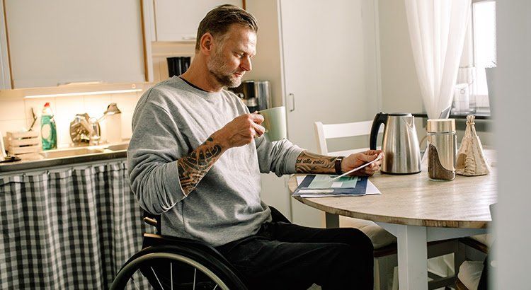 A man in a wheelchair is sitting at a table drinking coffee and looking at his phone.