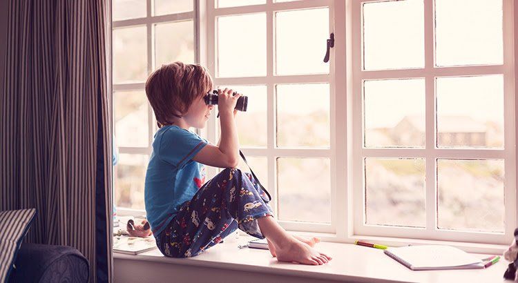 A young boy is sitting on a window sill looking through binoculars.