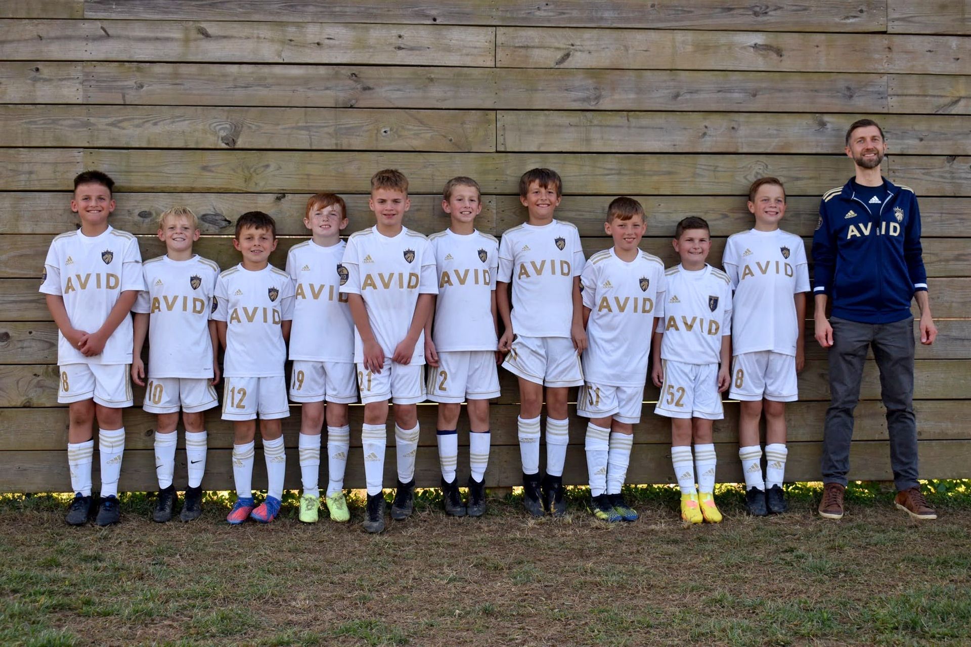 Youth soccer team in white uniforms standing in a row with a coach in front of a wooden wall