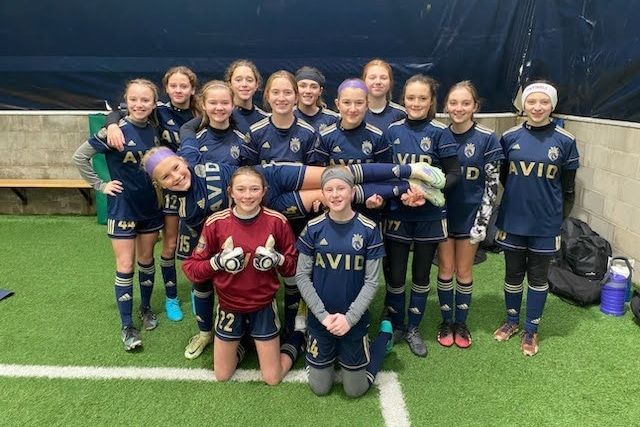 Youth soccer team posing on a turf field in navy blue uniforms, some holding trophies, smiling for a group photo
