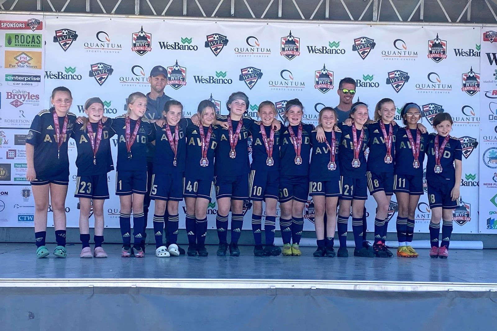 Soccer team posing on a stage with medals and trophies in front of a sponsor backdrop