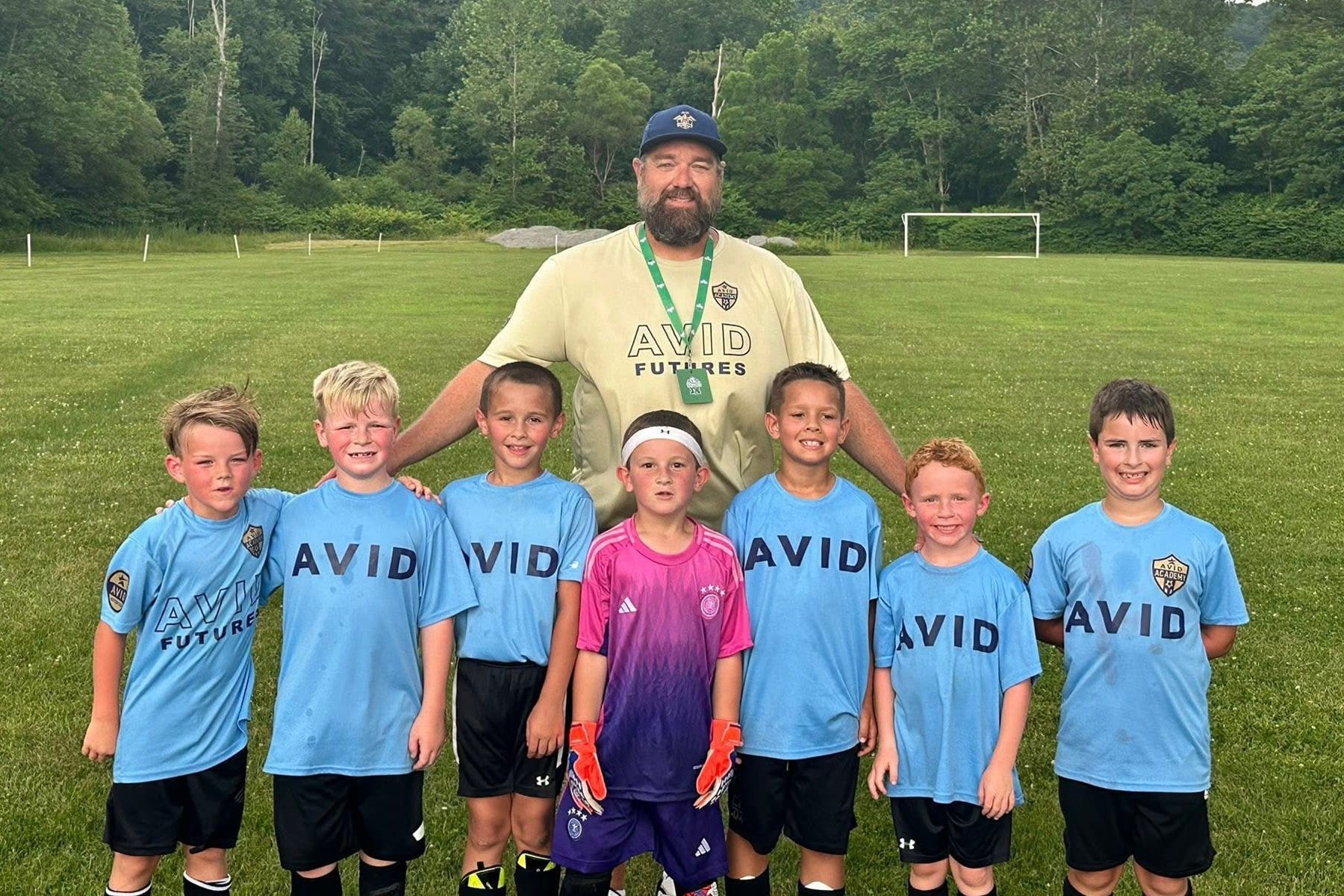 Youth soccer team posing with coach on a grassy field, wearing blue AVID shirts and one pink jersey.