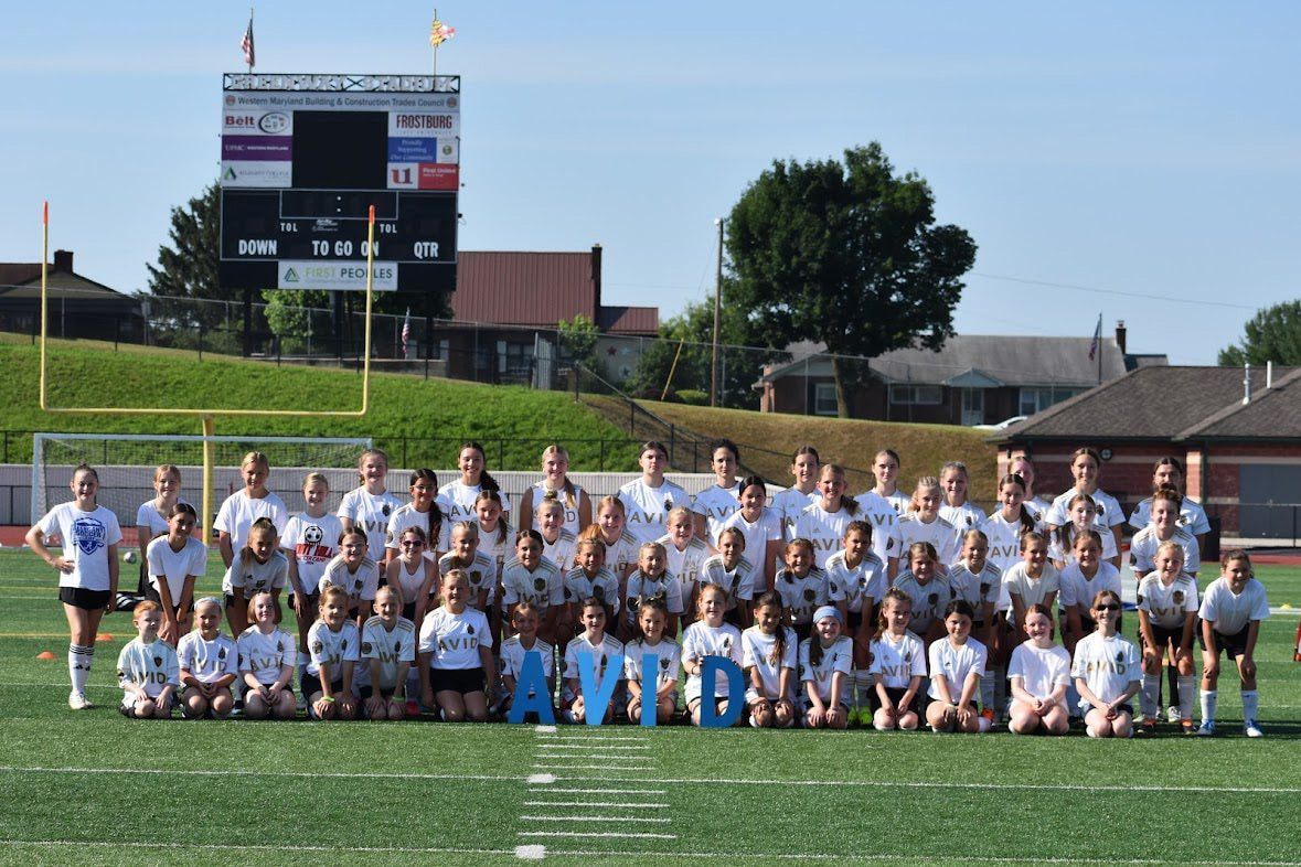 Large group posing on a sports field with cheerleading letters and a scoreboard in the background