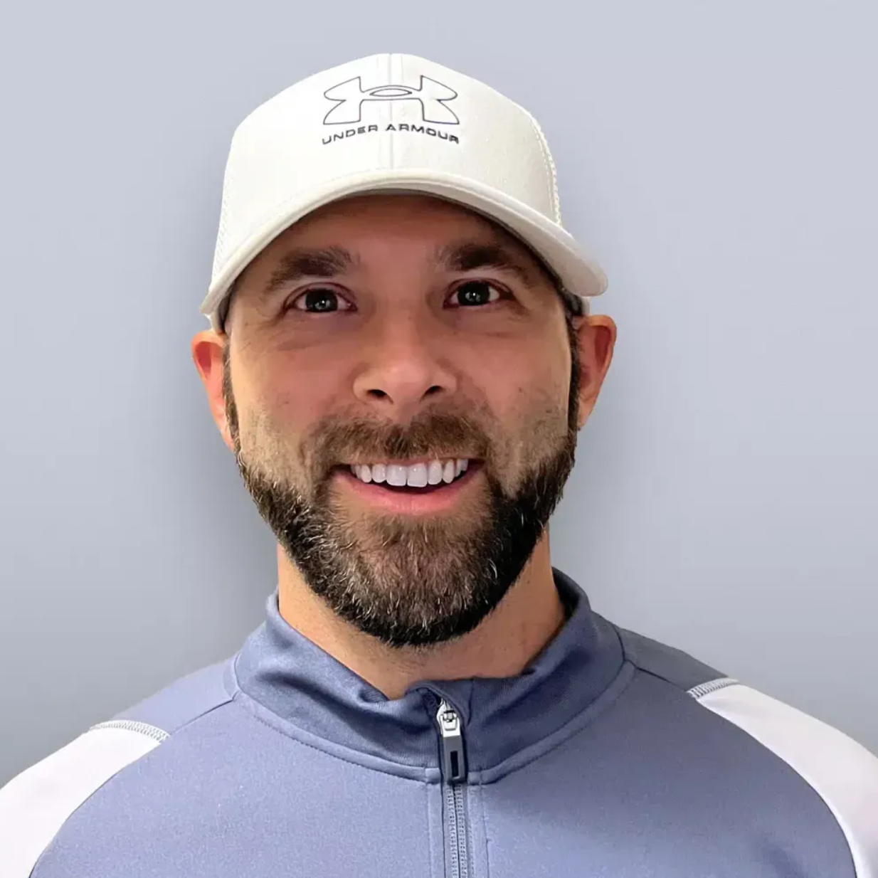 Smiling man in a white cap and light gray zip-up athletic shirt against a plain gray background