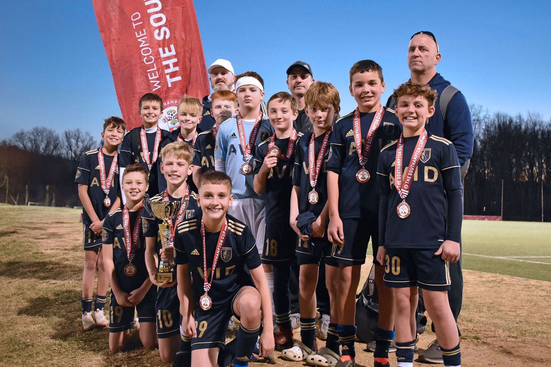 Youth soccer team posing with medals and a trophy on a field at dusk