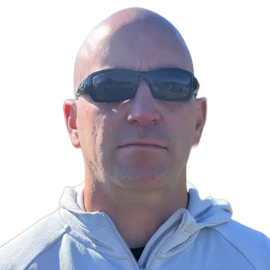 A bald man wearing sunglasses and a light hoodie stands outdoors in bright sunlight.