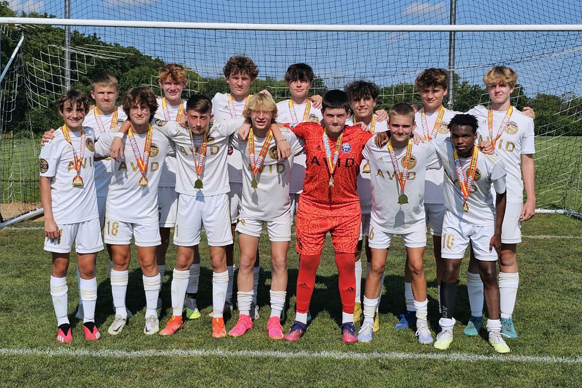 Soccer team posing on a field, with players in white uniforms and a goalkeeper in red in front of a goal