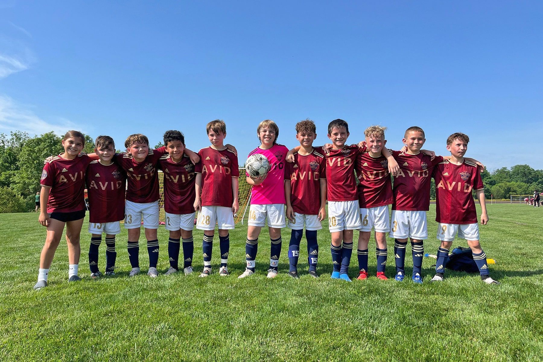 Youth soccer team posing on a grassy field under a clear blue sky, wearing maroon and pink jerseys