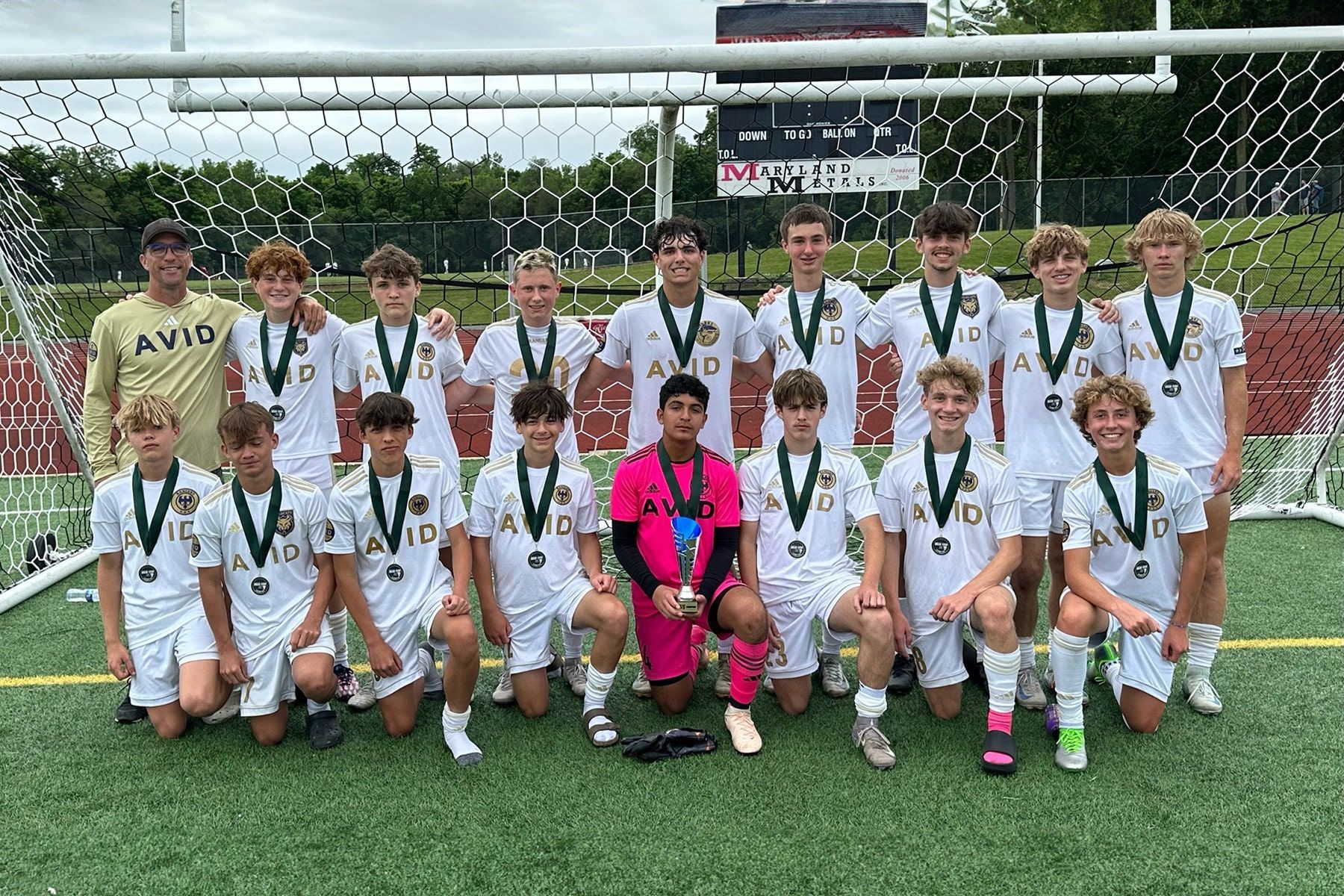 Soccer team posing on a field in front of a goal, wearing white uniforms and medals, with a trophy holder kneeling in front