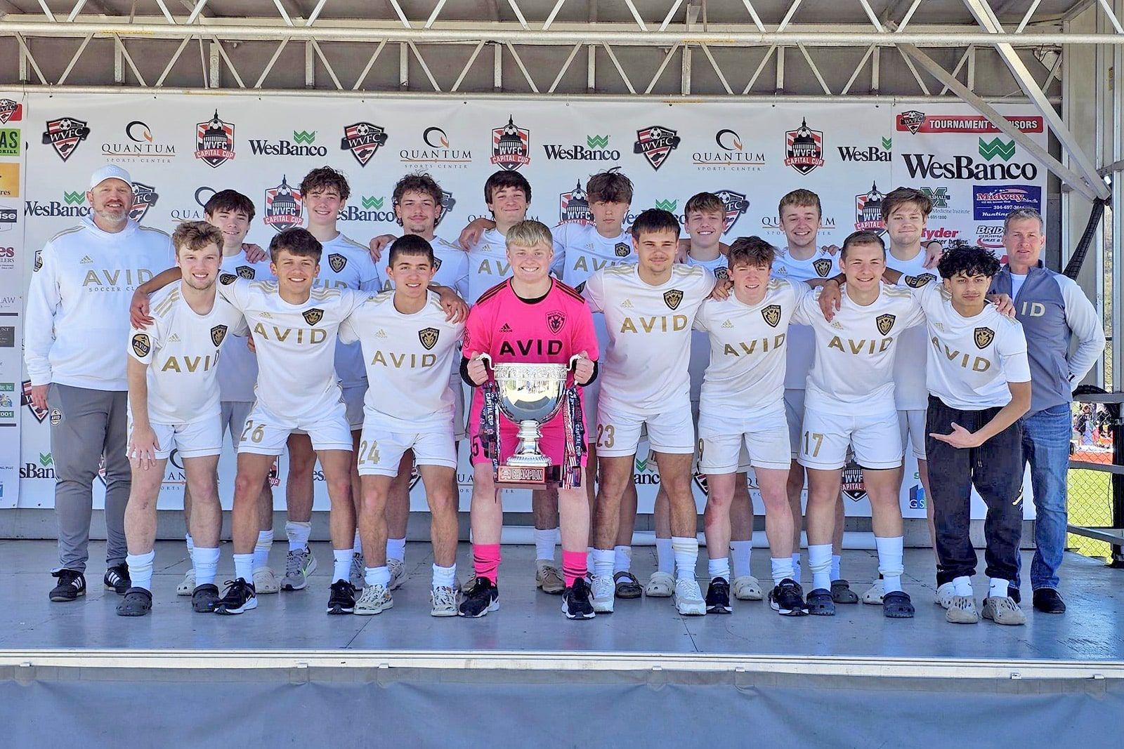 Soccer team posing on stage with trophy, white uniforms, and a goalkeeper in bright pink in the center