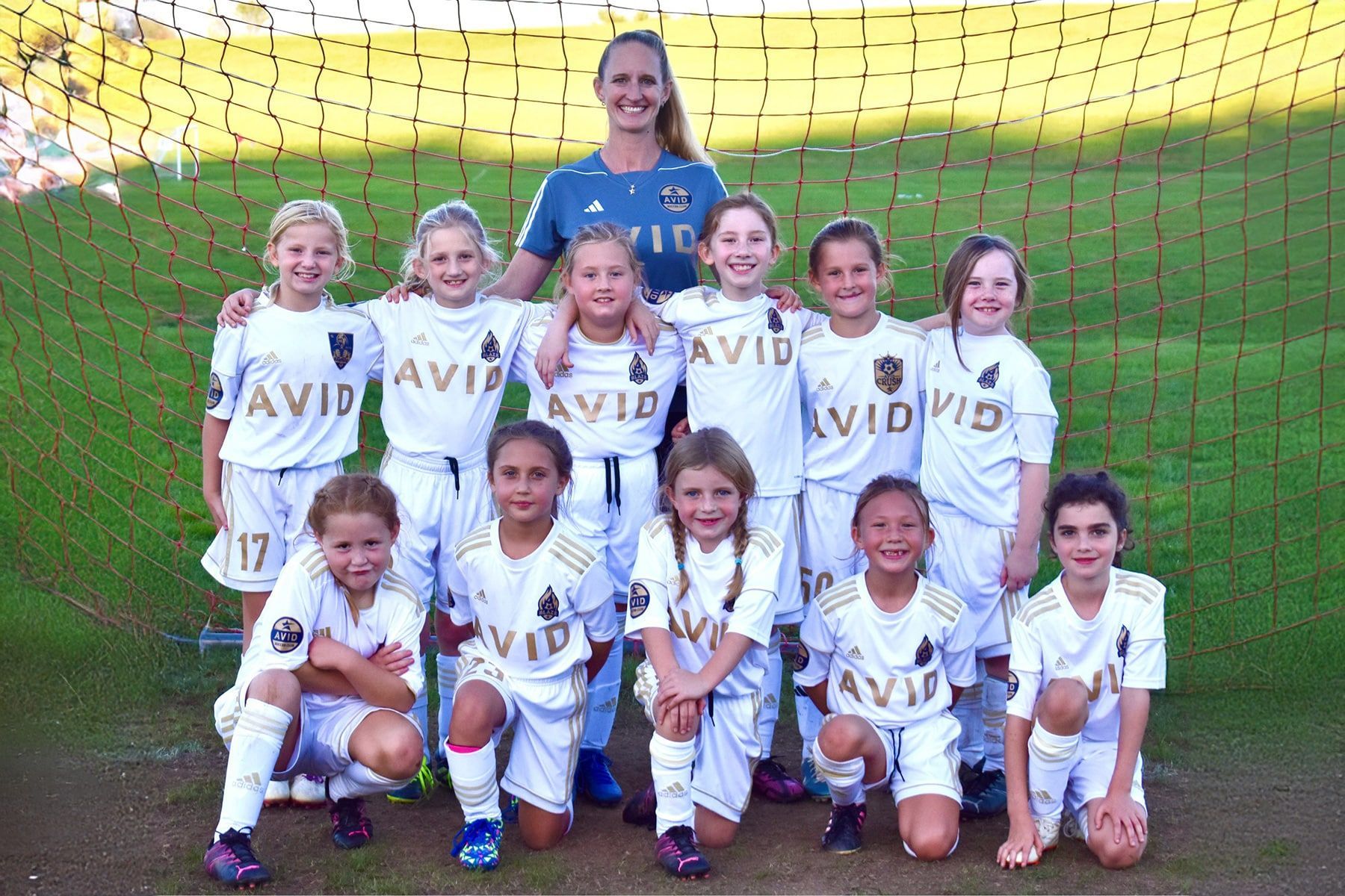 Youth soccer team posing with coach in front of a goal, wearing white uniforms and smiling.