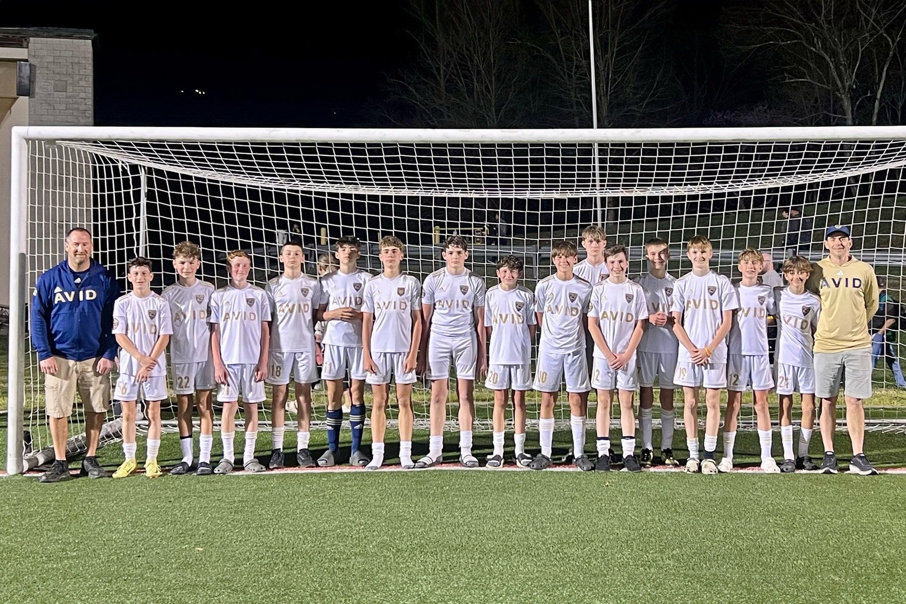 Soccer team lined up on a field at night in front of a goal, posing for a group photo.