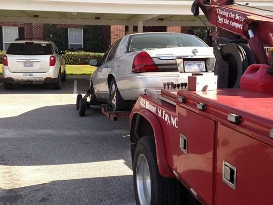 A silver car being towed by a red tow truck in a parking lot in Fayetteville, NC.