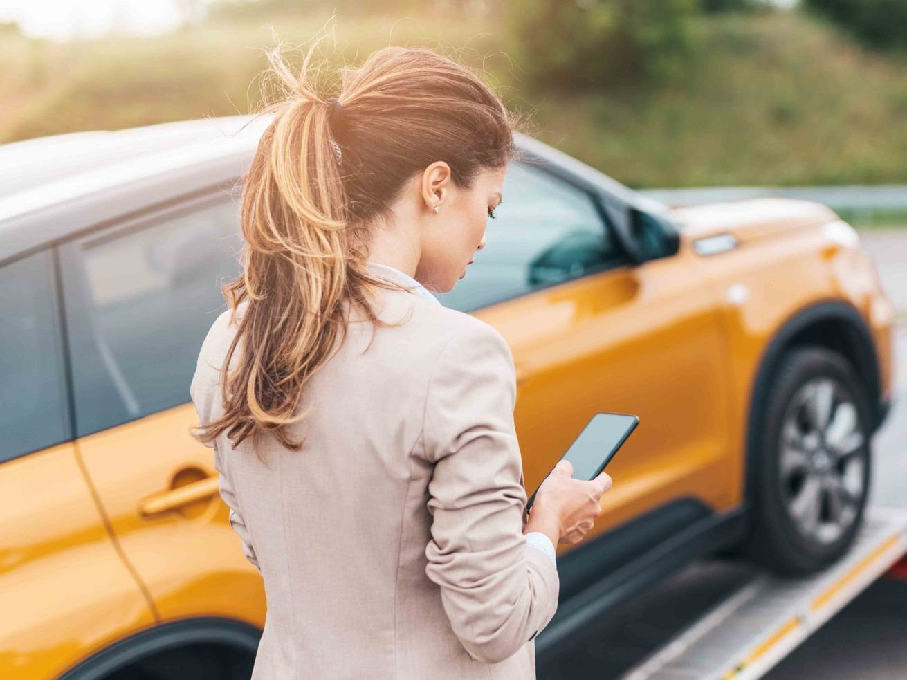 Woman in tan jacket, looking at phone, near a yellow SUV on a tow truck.