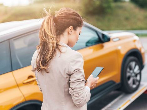 a woman is standing next to a broken down car and looking at her phone showing 24-hour roadside assistance Fayetteville NC.
