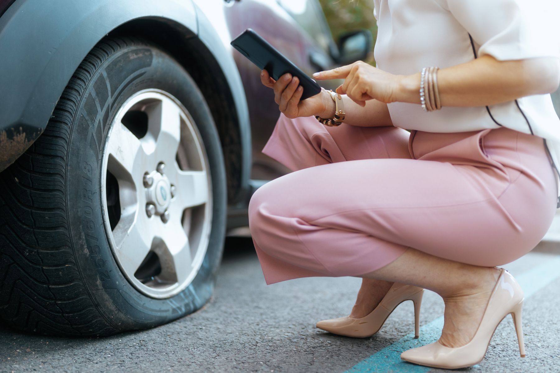 Woman crouching beside a flat car tire, holding a smartphone and keys on a roadside in Fayetteville, NC.