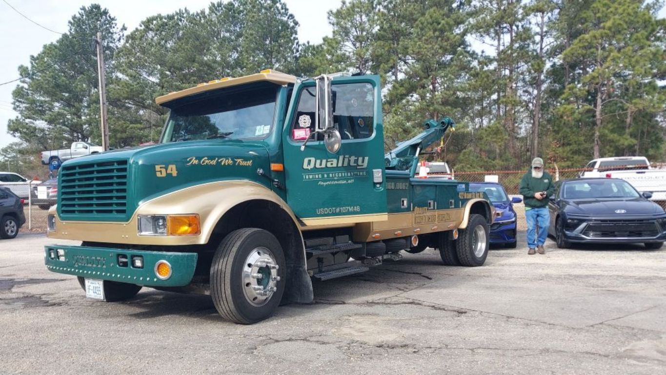 Woman with phone near an orange car loaded onto a tow truck in Fayetteville, NC.