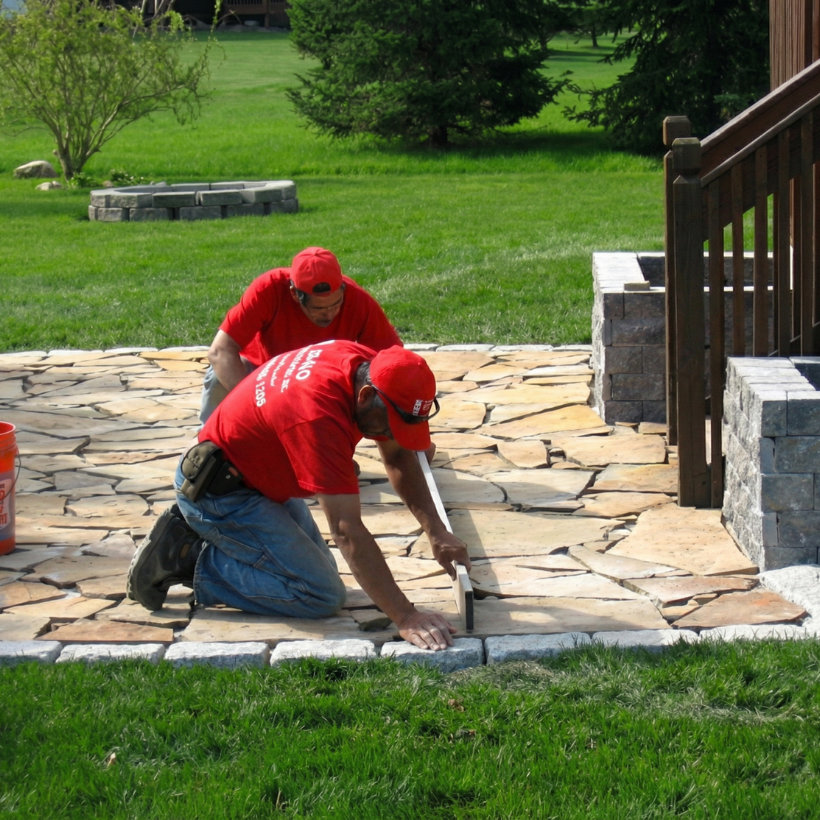 Two people from Bako Landscaping in red shirts and hats kneel while laying stone pavers on a patio next to wooden stairs.